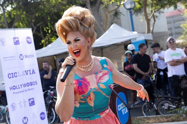 A drag queen in a colorful sleeveless dress and blonde bouffant wig speaking into a microphone at an outdoor event.